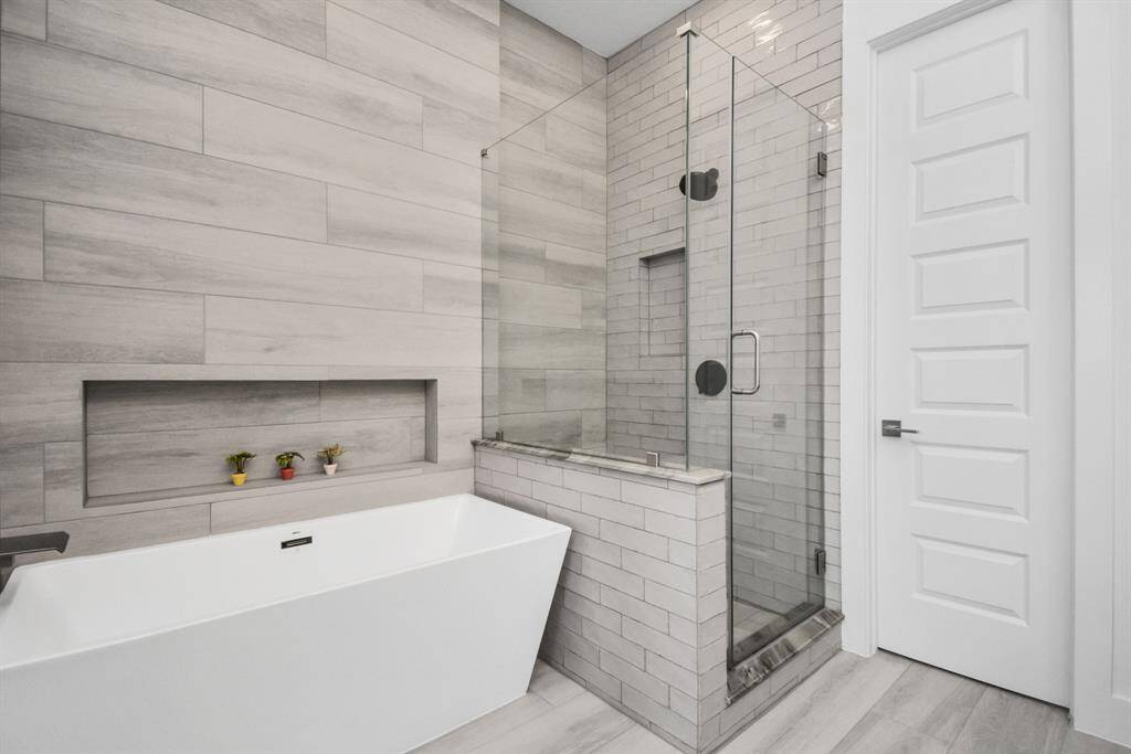 Modern bathroom featuring a sleek white bathtub, glass-enclosed shower, and stylish gray tile walls with built-in shelving.