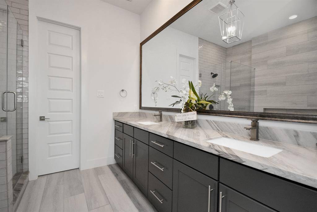 This modern bathroom features a spacious double vanity with sleek dark cabinetry and marble countertops. The large mirror, elegant light fixture, and glass-enclosed shower enhance the room's contemporary design.