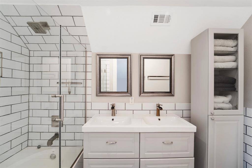 The primary bedroom features a beautifully appointed en-suite bathroom complete with a sleek tiled shower/tub combo with a glass door, and a double sink vanity topped with framed mirrors—offering both style and convenience.