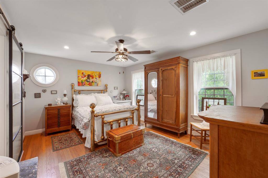 This spacious bedroom suite offers comfort and charm with multiple windows that bring in natural light, a ceiling fan paired with recessed lighting for a bright and airy feel, and rich hardwood floors that complete the polished look.