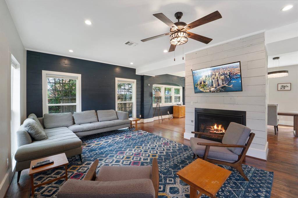 The inviting living room features rich hardwood floors, a cozy gas log fireplace, a ceiling fan for year-round comfort, and multiple windows that bathe the space in natural light.
