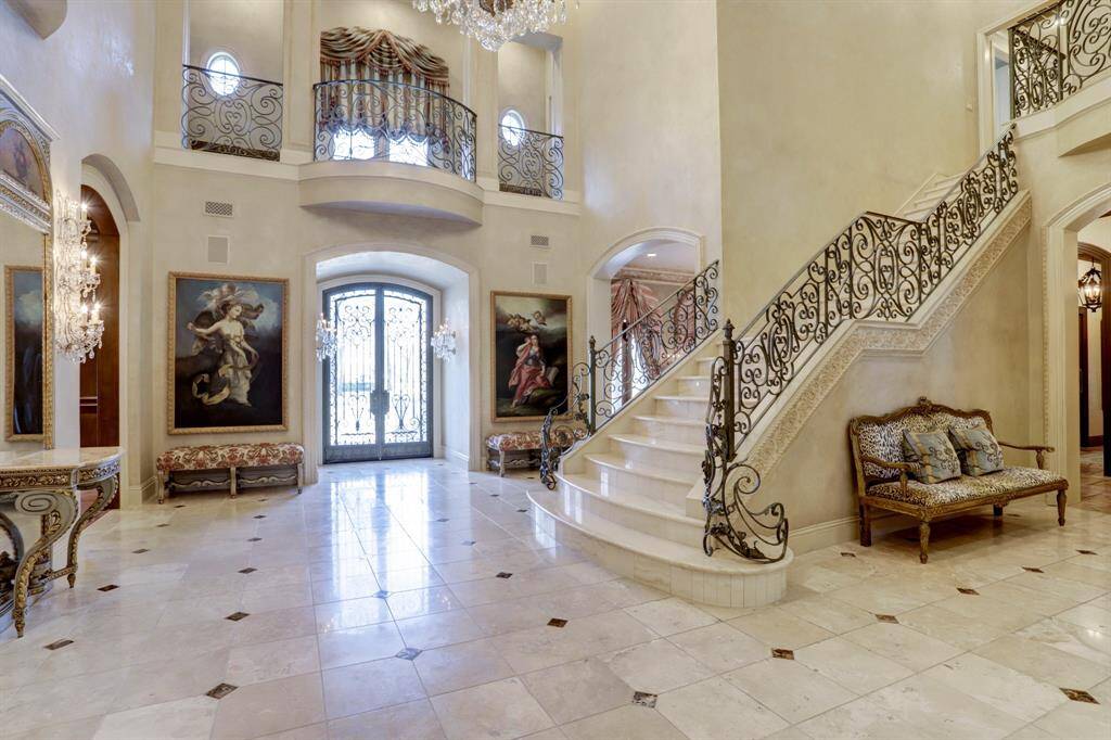 Grand foyer with soaring ceilings, extensive marble floors and staircase, and a stunning iron stair rail adorned with intricate details.