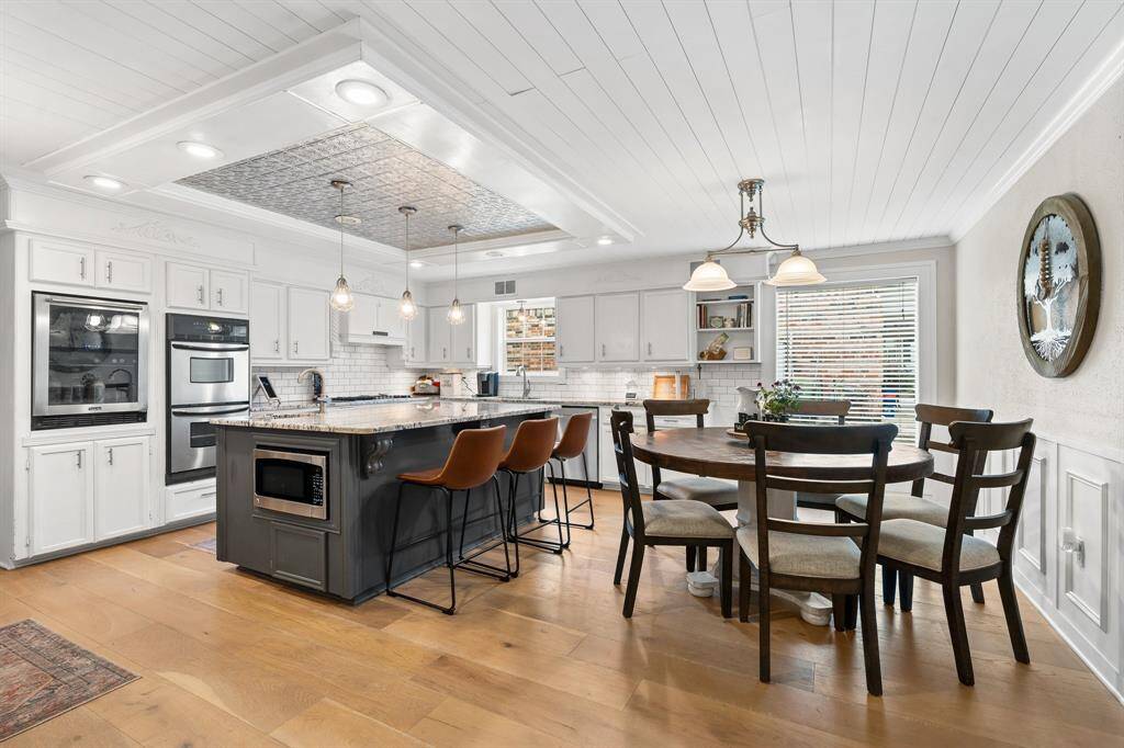 This modern kitchen features a spacious island with bar seating, stainless steel appliances, and a double oven. Stylish white cabinetry with elegant tile backsplash, and a cozy dining area.  The room is bright with natural light from a large window.