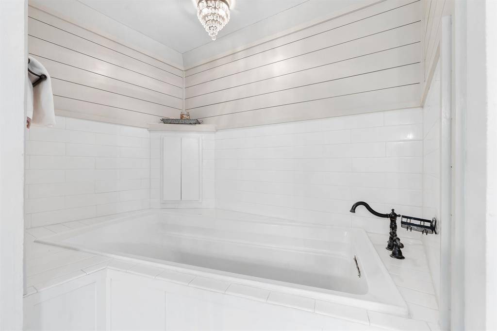 This photo features a modern bathroom with a large, white tiled soaking tub. The walls are accented with horizontal shiplap, and a stylish black faucet adds elegance. A small, decorative chandelier hangs above, providing a touch of luxury.
