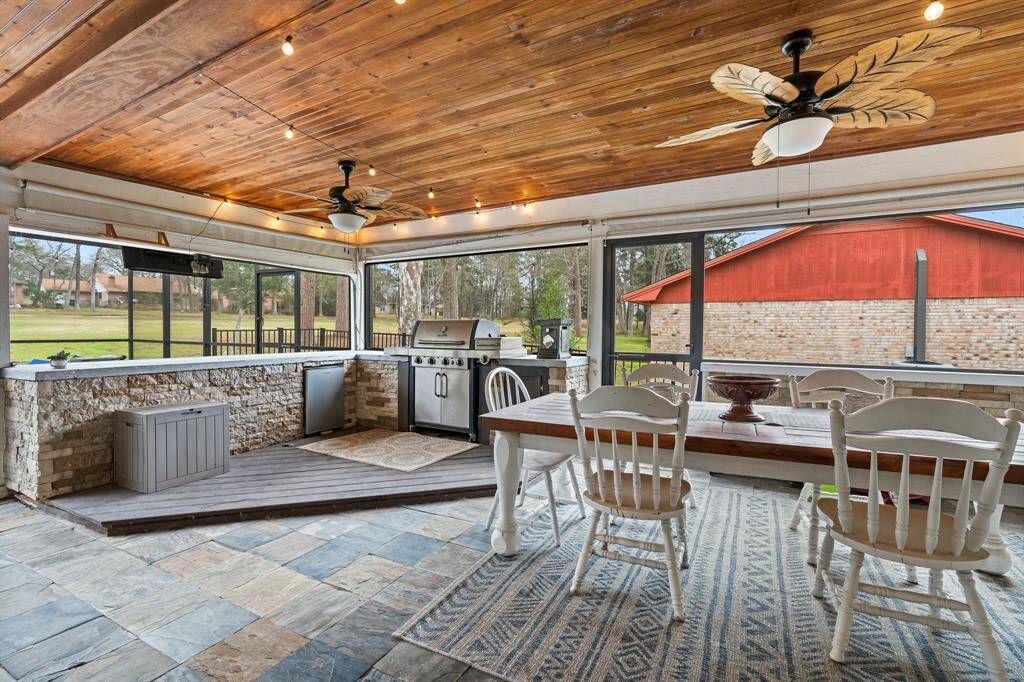 This inviting outdoor space features a spacious covered patio with a wooden ceiling. It includes outdoor kitchen, counter space, and a dining area perfect for entertaining. Large windows offer scenic views.