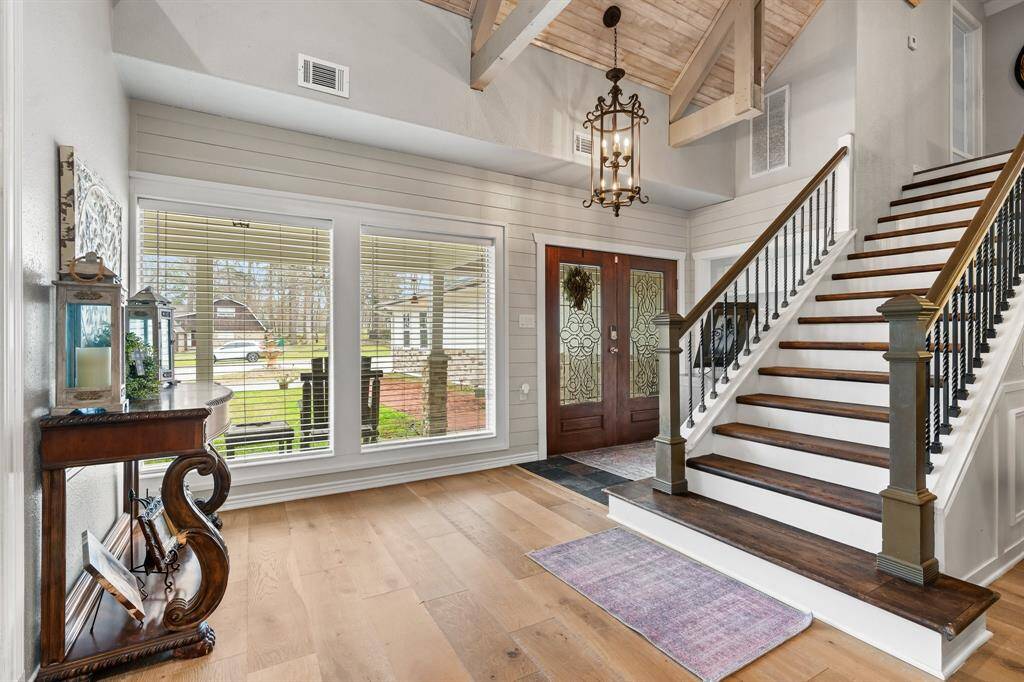 This inviting entryway features a stylish wooden staircase with elegant railings, a high vaulted ceiling with exposed beams, and a large chandelier. Natural light pours through the expansive windows, highlighting the warm wood floors and decorative accents. A glass-paneled front door adds to the welcoming atmosphere.