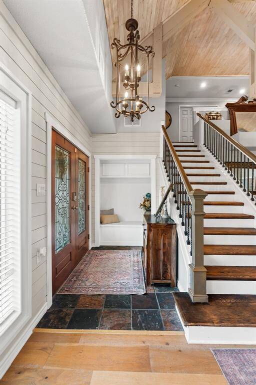 This inviting entryway features a warm, rustic design with wooden accents and a high ceiling. A grand staircase with dark railings leads to the upper level, while a stylish chandelier adds elegance. The space includes a cozy bench nook and decorative elements, creating a welcoming atmosphere.