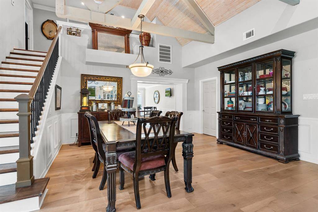 This inviting dining area features high ceilings with exposed beams and elegant wainscoting. The room is well-lit by a central pendant light and offers a view of a staircase and upper loft area, adding to the open, airy feel.