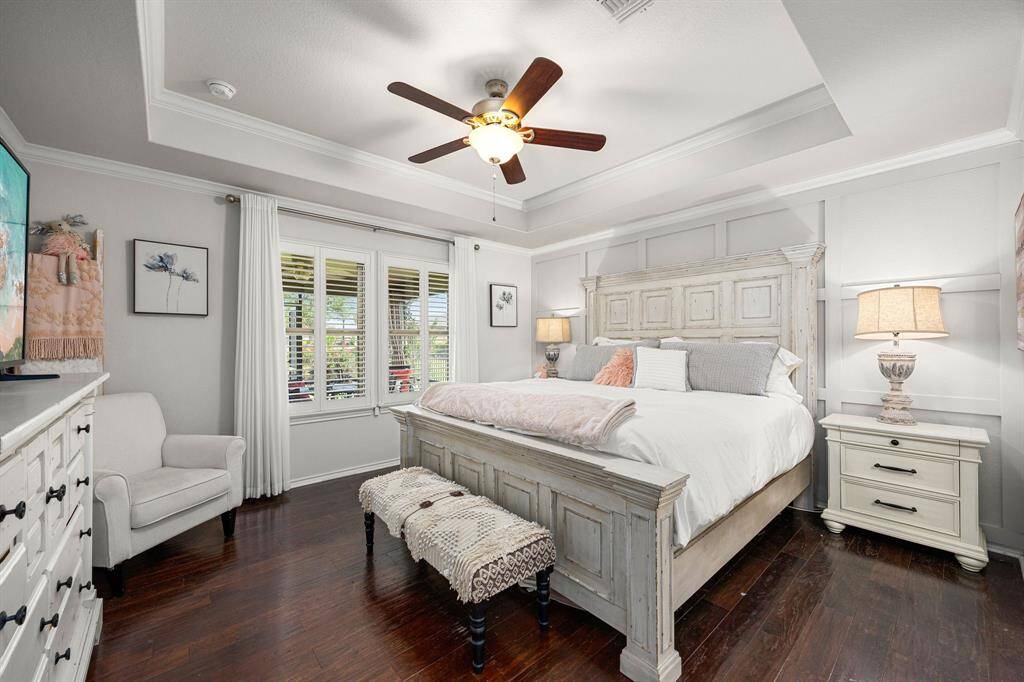 The Primary bedroom has elegant tray ceilings, a ceiling fan, and large windows with Plantation shutters overlooking the screened in patio. Wooden flooring and gray wainscoted wall offer soft, neutral-toned decor that creates a warm and relaxing atmosphere.