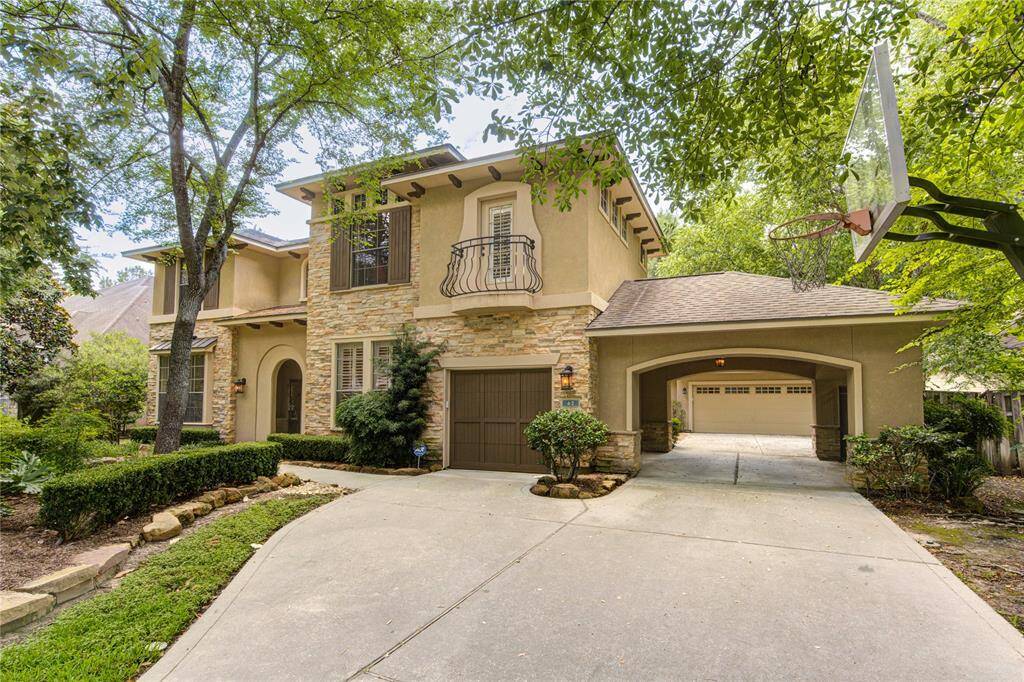 This elegant two-story home features a stone and stucco exterior with a charming balcony and arched entryway. The spacious driveway leads to a double garage, and the lush landscaping includes mature trees and well-manicured hedges, creating a welcoming and private atmosphere.