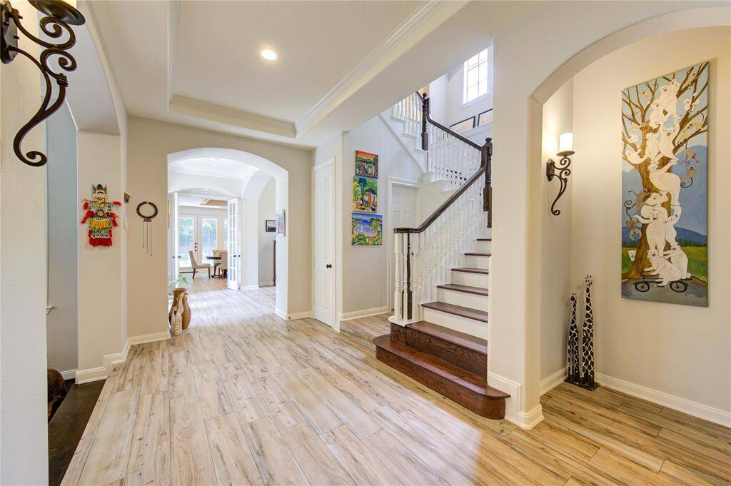 This entryway features elegant wood flooring and high ceilings, with a grand staircase leading to the upper level. Bright artwork and decorative sconces add charm, while an archway opens to a sunlit dining area.