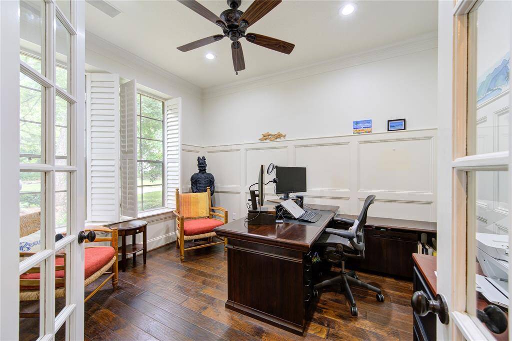 Bright home office with large windows, plantation shutters, and hardwood floors. Features a ceiling fan, built-in desk, and comfortable seating. Ideal for productivity and relaxation.