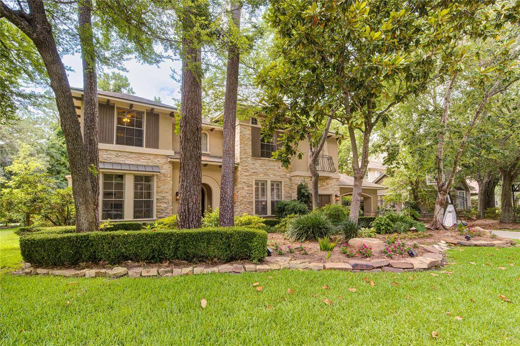 This photo features a charming two-story home with a stone and stucco exterior, nestled among mature trees and lush landscaping. The well-maintained front yard includes a manicured lawn, flower beds, and a stone-lined path, offering great curb appeal in a serene setting.