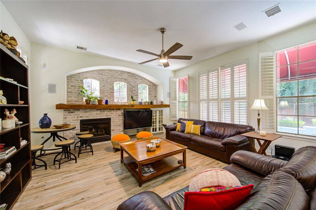This inviting living room features a cozy fireplace with a brick accent wall, large windows with plantation shutters for ample natural light, and sleek hardwood floors. The space is furnished with comfortable leather seating and a modern ceiling fan, creating a warm and welcoming atmosphere.