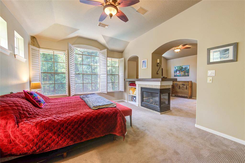 This spacious bedroom features large bay windows with shutters, a cozy fireplace, and a ceiling fan. The room is carpeted and includes an archway leading to an adjacent area, offering a versatile space perfect for relaxation or additional furnishings.