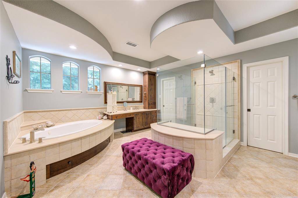 This spacious bathroom features a luxurious design with a corner bathtub, a large glass-enclosed shower, and elegant tile work. It includes a stylish vanity with ample storage and three windows for natural light. The room is accented by a unique ceiling design and a plush purple bench.