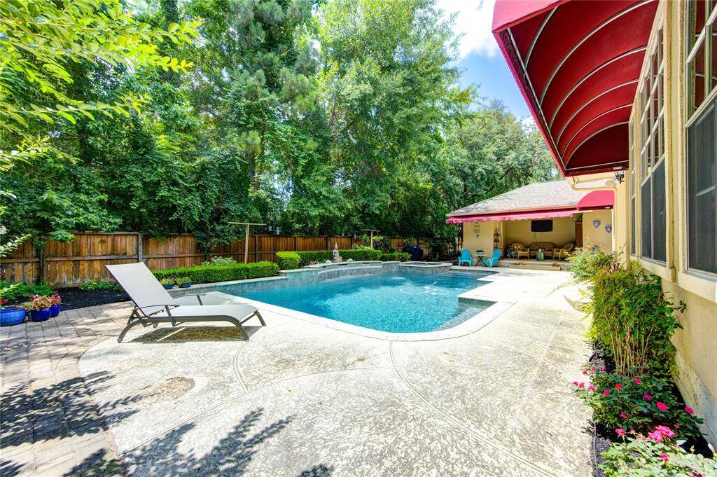 This photo showcases a serene backyard with a sparkling pool, surrounded by lush greenery and a wooden fence. The patio features comfortable seating under a shaded canopy, providing an inviting space for relaxation and entertaining.