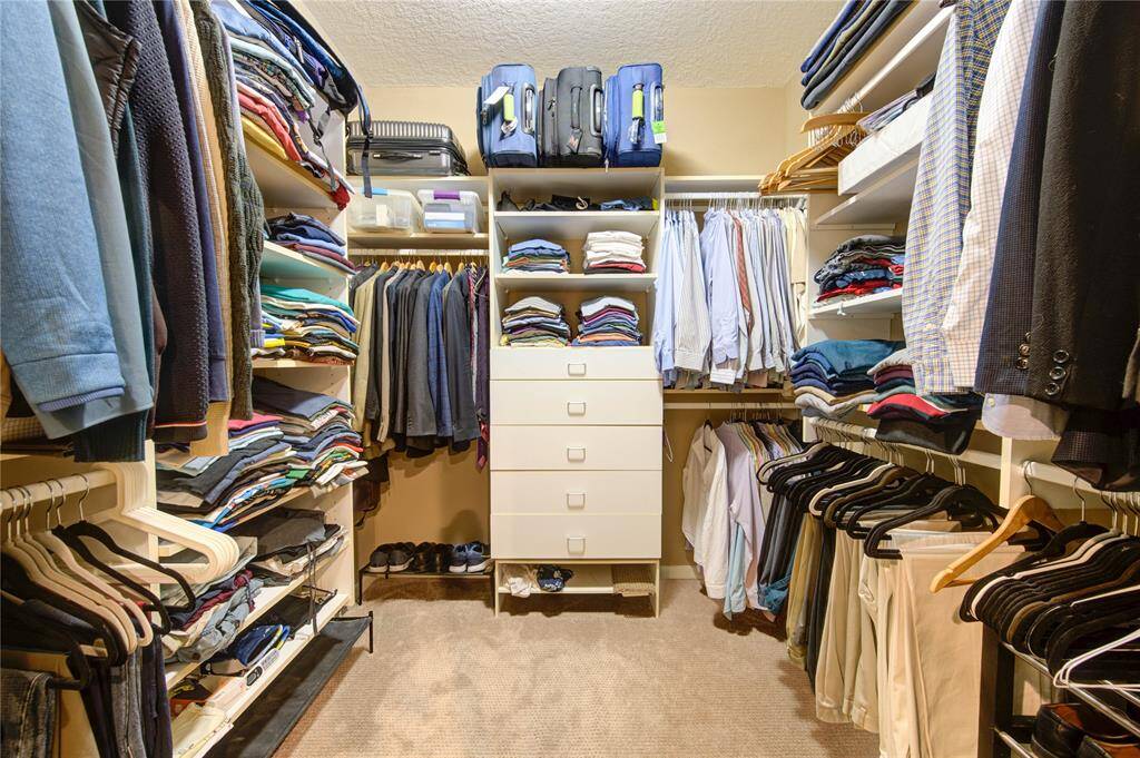 This spacious walk-in closet features ample shelving, hanging space, and drawers, perfect for organizing clothing and accessories. The neutral carpet and lighting create a cozy atmosphere. One of the 2   primary  bedroom closets
