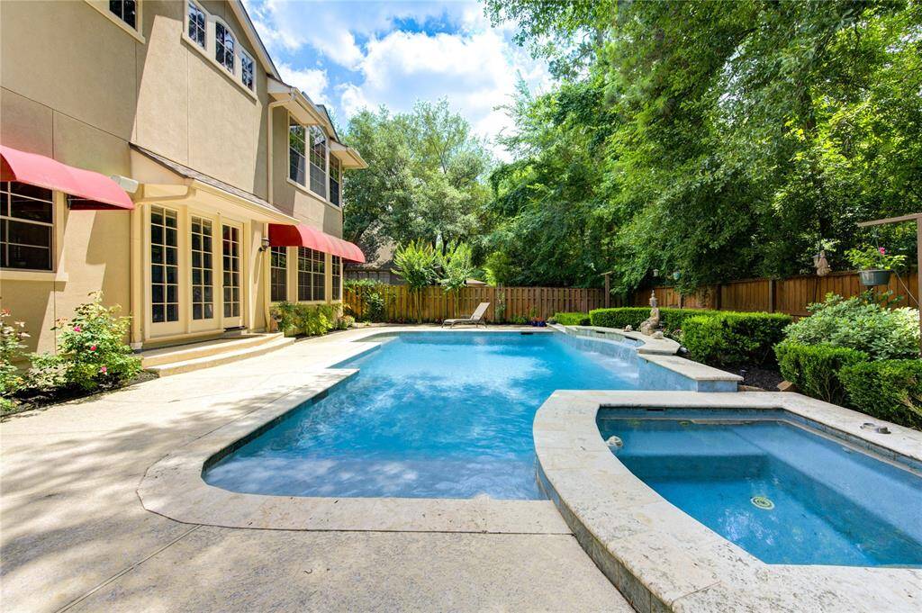 This backyard features a spacious pool and spa, surrounded by lush greenery and a privacy fence. The home’s exterior has large windows and charming red awnings, offering a welcoming and serene outdoor retreat.