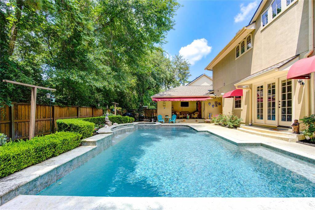 This photo showcases a charming backyard with a spacious pool, surrounded by lush greenery and a wooden fence. The house features elegant French doors and red awnings, offering a cozy patio area perfect for relaxation and entertaining.
