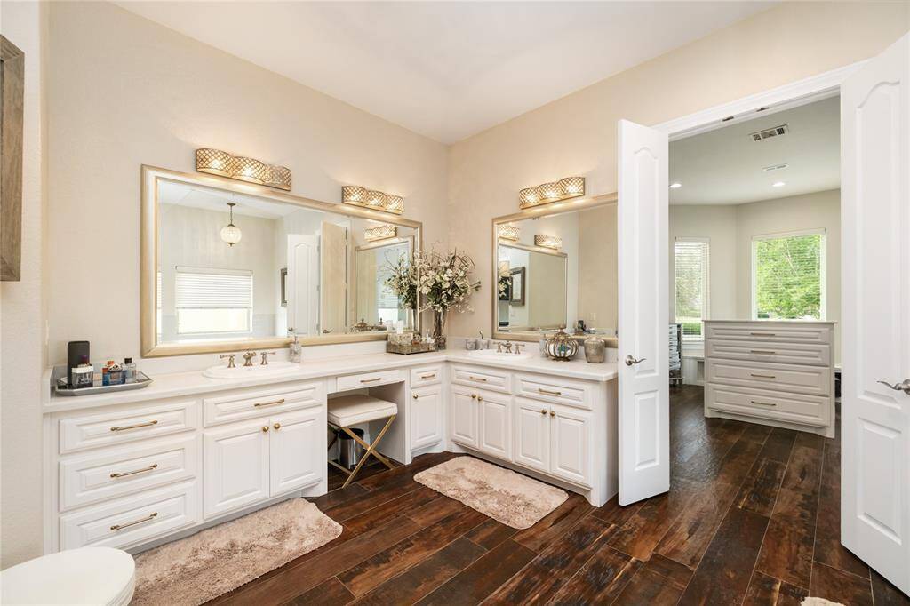 The vanity is a stunning focal point, featuring crisp white cabinetry adorned with gold hardware and elegant framed mirrors. Gold-finished light fixtures add a warm, refined glow, while ample cabinet storage keeps the space beautifully functional. The contrast between the white cabinetry and gold accents creates a timelessly sophisticated aesthetic.