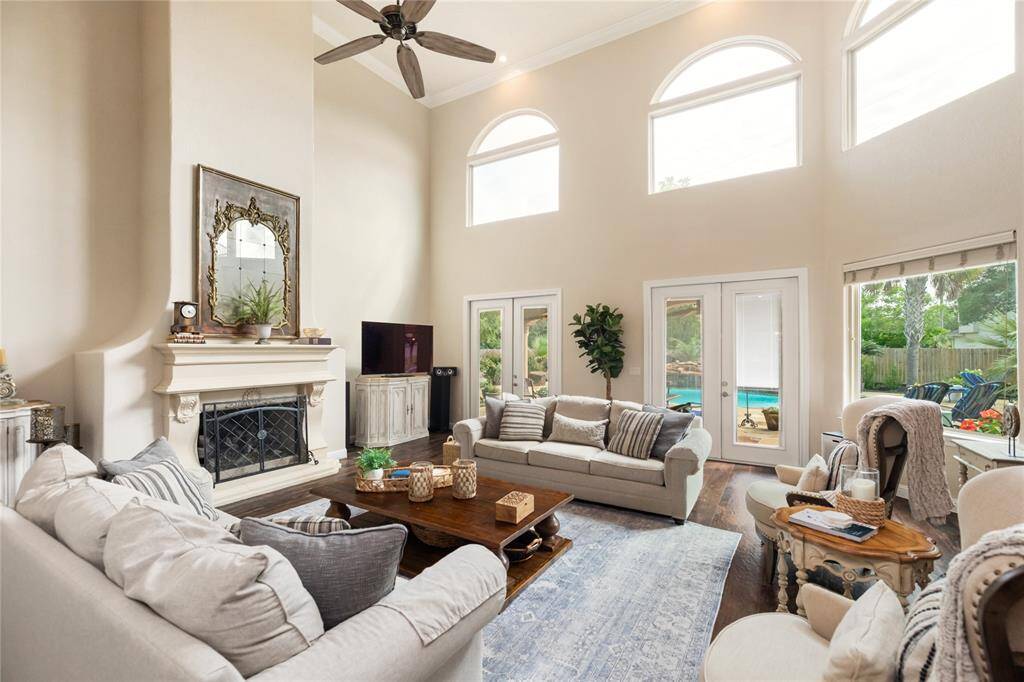 This breathtaking living room impresses with soaring ceilings, a stately fireplace framed by an elegant mantle, and grand arched windows that bathe the space in natural light. French doors open directly to the backyard oasis, while crown molding and ceiling fans add classic and functional detail.