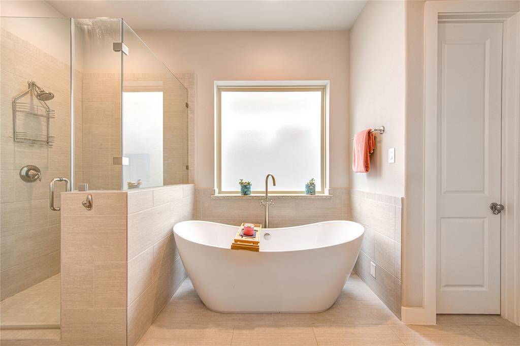 Luxurious and spa-inspired, the primary bathroom showcases a freestanding soaking tub perfectly positioned beneath a large window for natural light and serene views. Adjacent to the tub is a spacious frameless glass walk-in shower with sleek tile surround and a built-in bench.