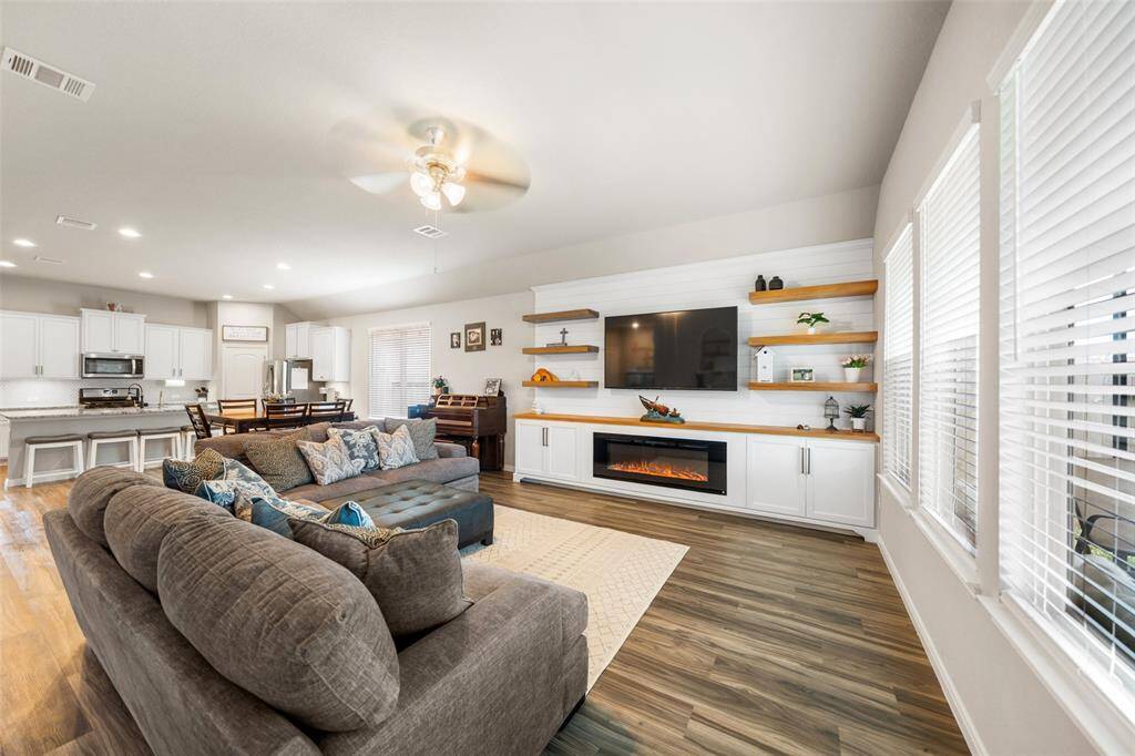 This photo showcases a spacious, open-concept living area. One of a kind custom elegant built-in entertainment center with an electric fireplace and large windows providing ample natural light. The kitchen is visible in the background creating a seamless flow between living and dining spaces.