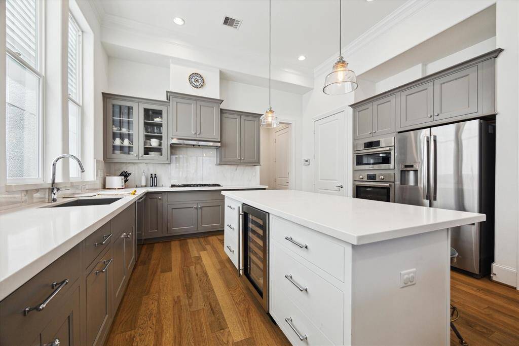The kitchen features high ceilings, sleek gray cabinets, a spacious island with seating and pendant lighting, stainless steel appliances, and ample natural light from large windows, creating a modern and inviting chef's space.