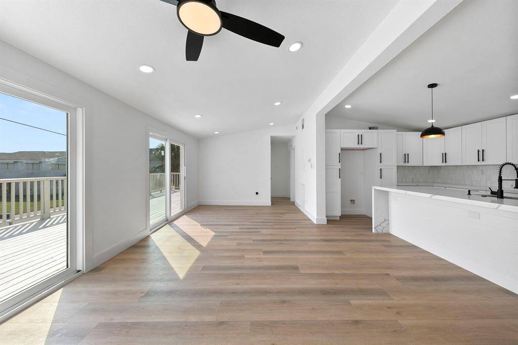This bright, open-concept living space features modern wood flooring, a ceiling fan, and ample natural light from large sliding glass doors leading to a deck. The adjacent kitchen boasts sleek white cabinetry, black fixtures, and a stylish pendant light.