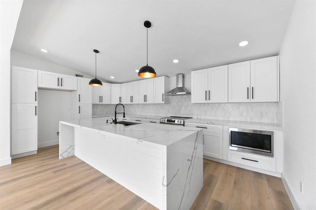 This modern kitchen features a spacious layout with white cabinetry, sleek black hardware, and marble countertops. It includes stainless steel appliances, a large island with a built-in sink, and stylish pendant lighting. The light wood flooring adds warmth to the bright, open space.