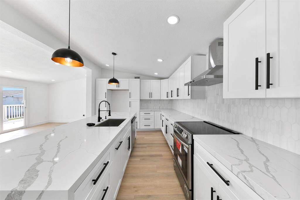 This modern kitchen features sleek white cabinetry, marble-patterned countertops, and a stylish backsplash. It includes a large island with a built-in sink and pendant lighting, along with stainless steel appliances. The space is bright and open, ideal for entertaining.