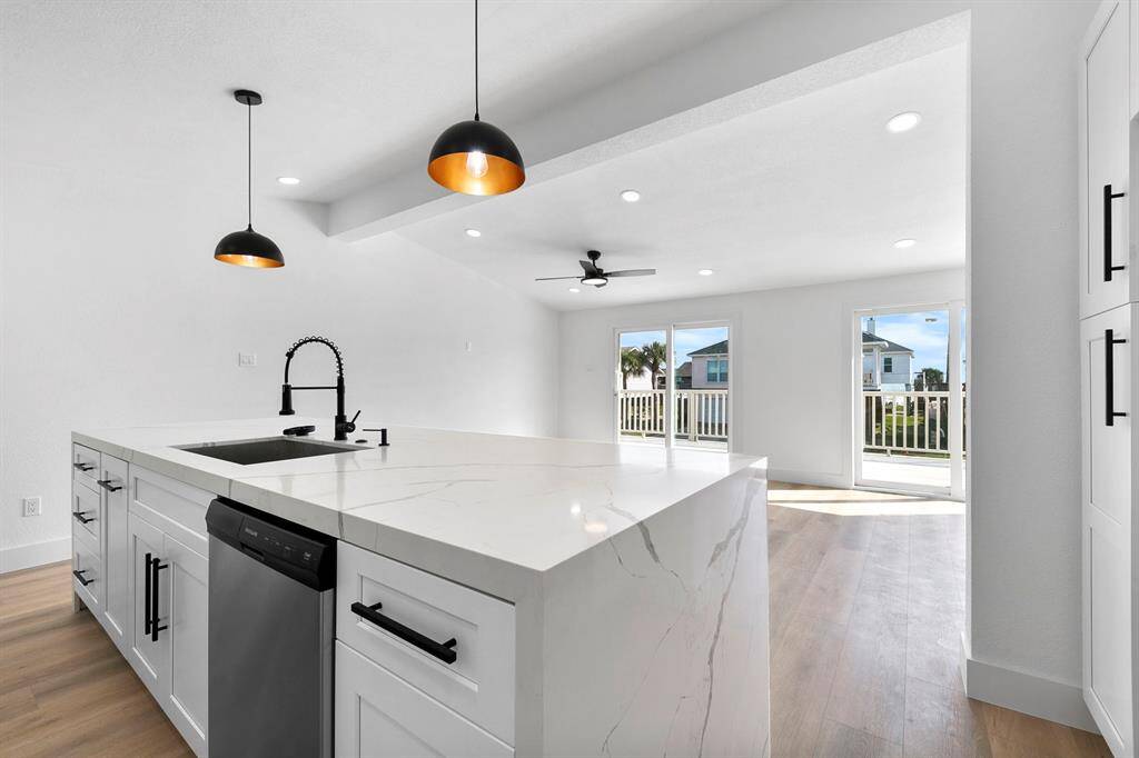 This modern kitchen features a sleek white island with a marble countertop, pendant lighting, and a built-in dishwasher. It opens to a bright living area with large windows and a view, offering a spacious and airy feel.