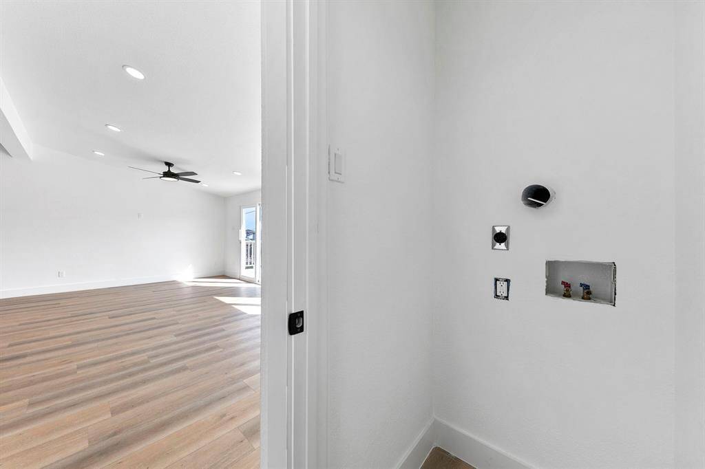 This photo showcases a bright, open living area with light wood flooring and recessed lighting. There is a ceiling fan and large windows that allow natural light to flood the space. A partial view of a utility area with plumbing and electrical outlets suggests washer and dryer hookups are nearby.
