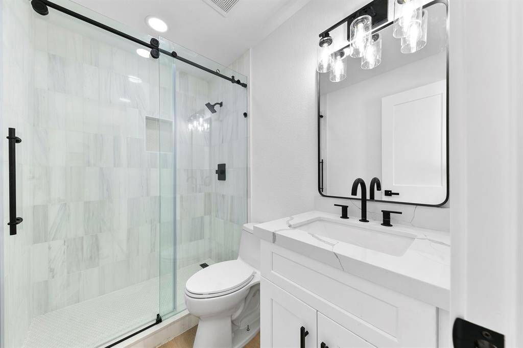 This bathroom features a sleek, modern design with a glass-enclosed shower, white tile, and black hardware. The vanity includes a marble countertop and contemporary fixtures, complemented by a stylish mirror and lighting.