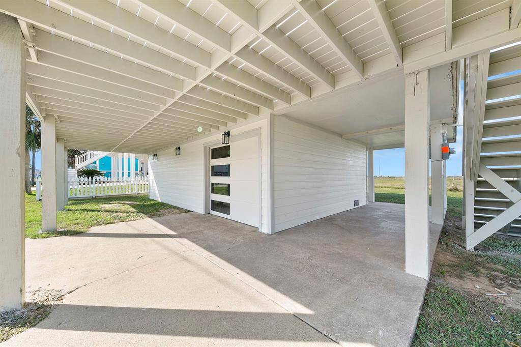 This photo shows an under-house parking area with a concrete floor, white siding, and an adjacent staircase leading up. It offers sheltered parking and storage space, with a view of a grassy yard and a white picket fence nearby.