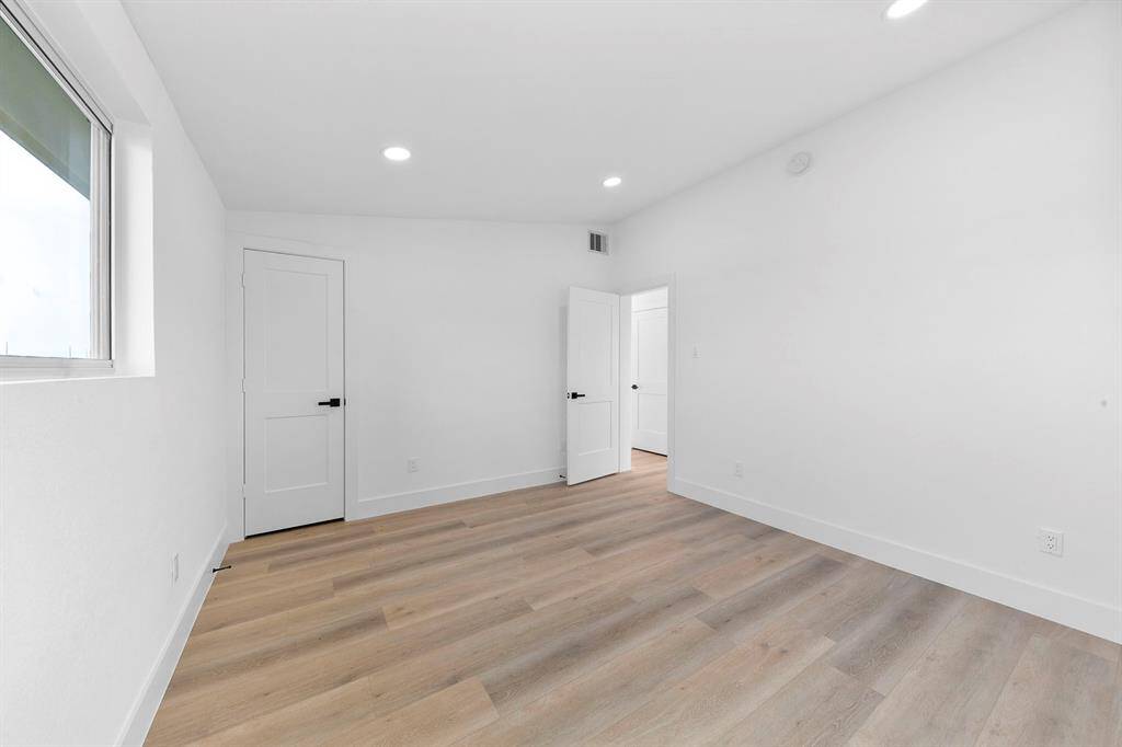Bright, minimalist room with light wood flooring, white walls, and recessed lighting. Features a window for natural light and a modern door. Ideal for a bedroom or office space.