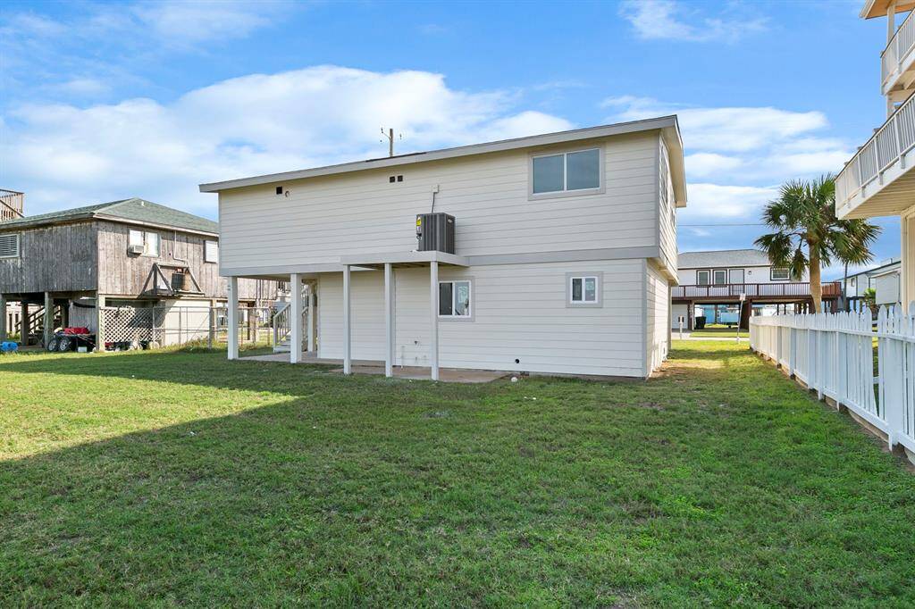 This photo shows the back of a raised, two-story coastal home with beige siding. It features an open, grassy backyard and is surrounded by neighboring elevated houses, offering a view of the nearby water.