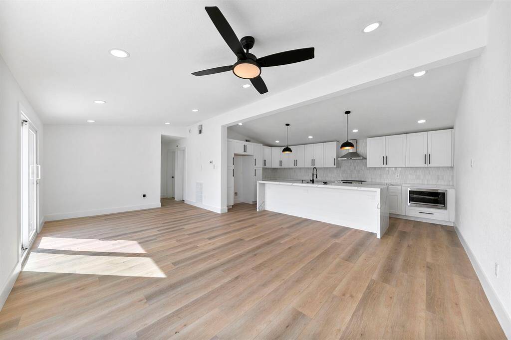 This photo showcases a bright, open-concept living space with modern finishes. It features light wood flooring, a sleek ceiling fan, recessed lighting, and a spacious kitchen with white cabinetry, a large island, and pendant lights. Natural light streams in through glass doors, enhancing the airy feel.