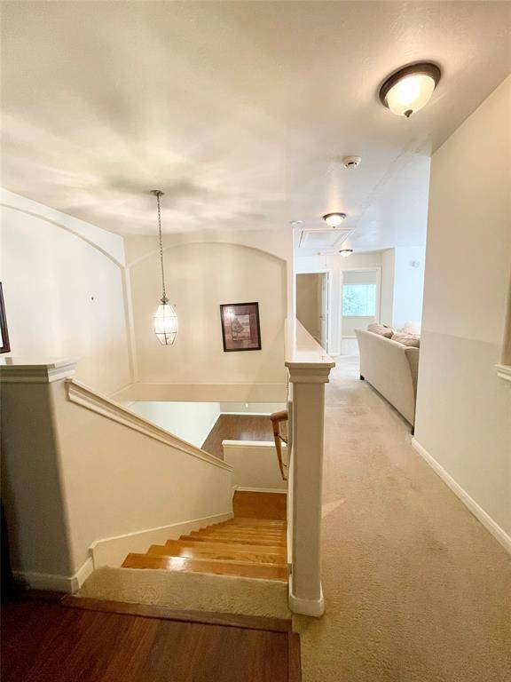 Hallway with carpeted flooring leading to a cozy living area. There are stairs with a wooden finish and an elegant hanging light fixture. The space appears open and inviting, with neutral wall colors and simple decor.