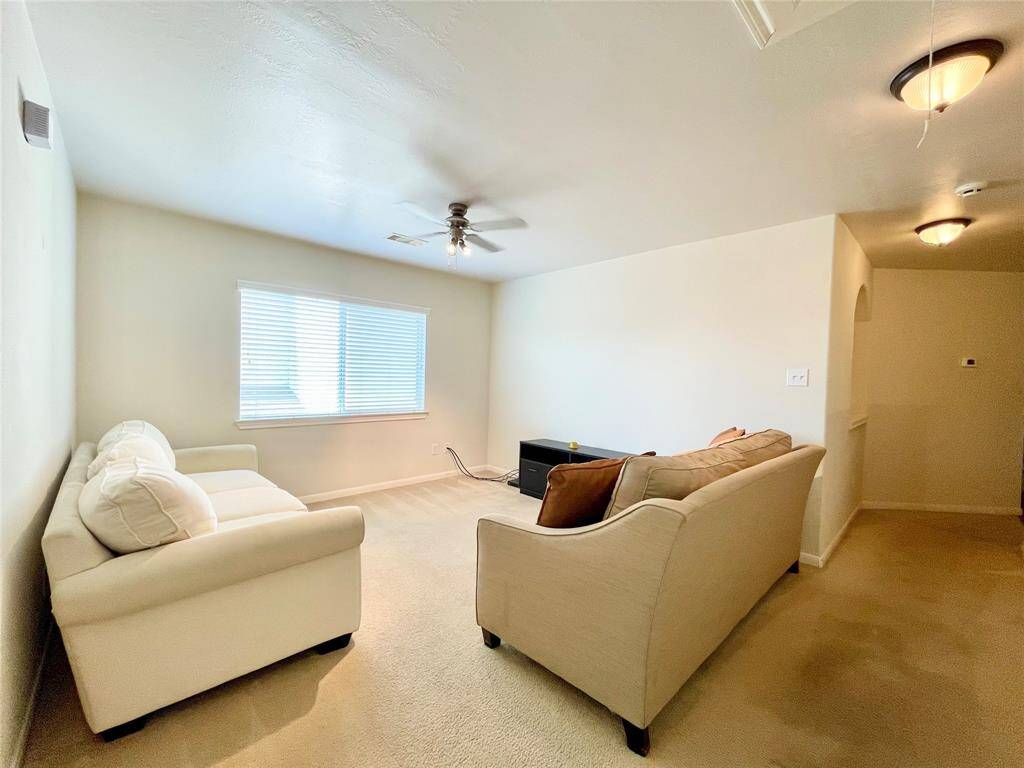 Bright game room with neutral walls and carpeting, featuring a ceiling fan, and a large window allowing natural light. Cozy and inviting space.