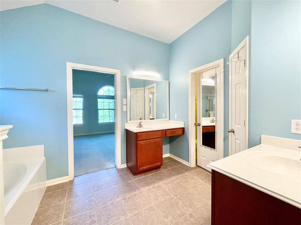 Bright master bathroom with blue walls, featuring dual sinks, a large mirror, and a bathtub. A door leads to a spacious  main bedroom.