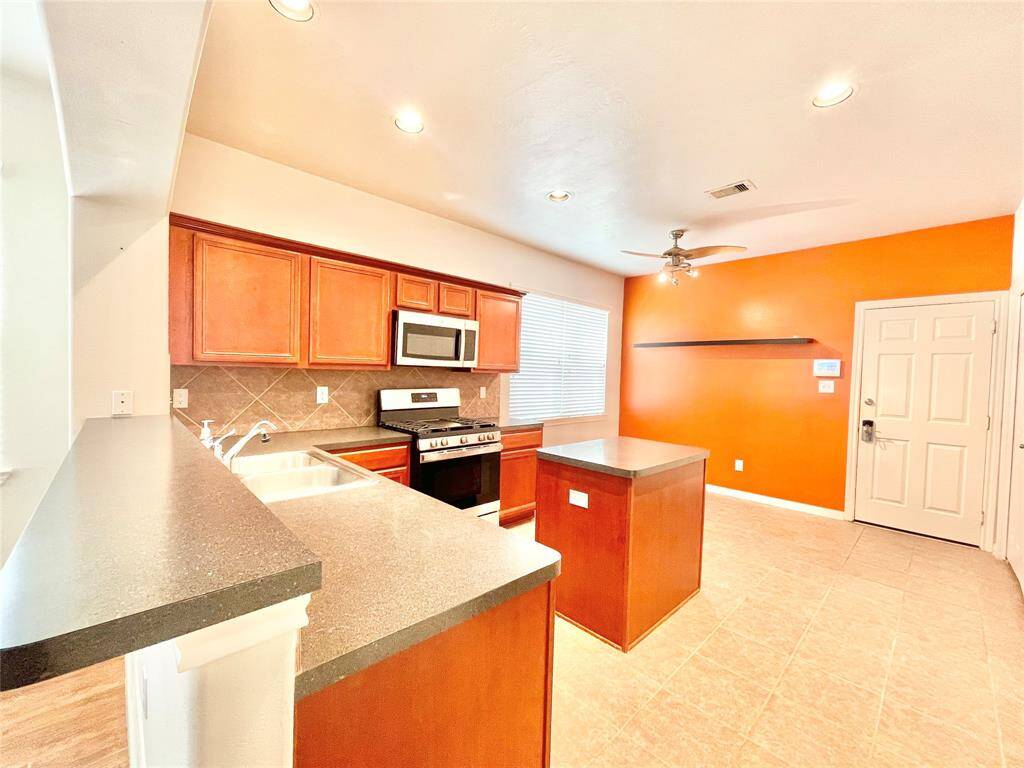 Bright kitchen with warm wooden cabinets, stainless steel appliances, and a central island. The space includes a double sink, contrasting countertops, and an eye-catching orange accent wall, creating a cozy atmosphere.