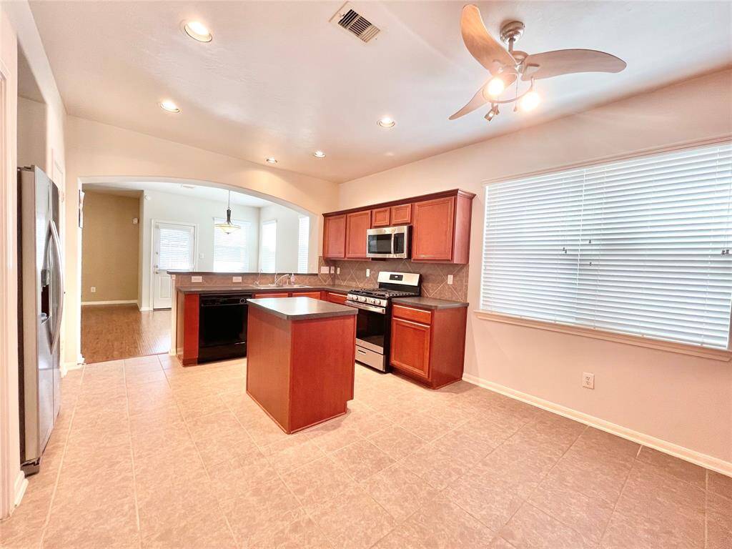This kitchen features modern stainless steel appliances, ample wooden cabinetry, a central island, and a ceiling fan. Natural light fills the space through large windows, and it opens up to an adjoining dining area.
