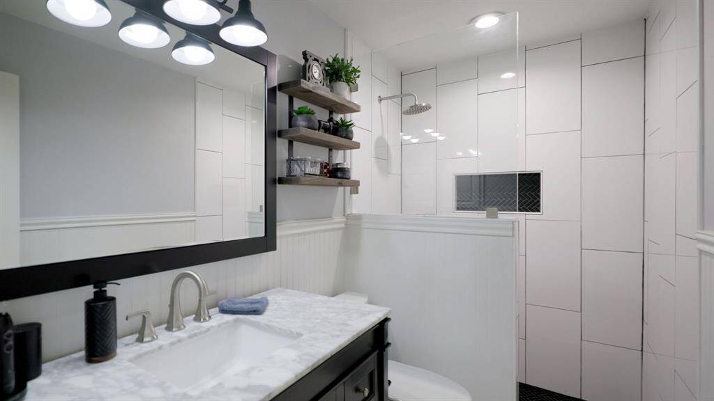This stylishly updated guest bathroom features a custom sink vanity with modern fixtures, a framed mirror with designer lighting, a sleek walk-in shower with large-scale tile, and a rainfall showerhead.