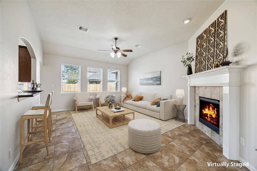 Spacious living area features tile flooring, a cozy fireplace, and neutral walls, offering a warm and inviting atmosphere. A ceiling fan adds comfort, while the open layout connects seamlessly to the kitchen.