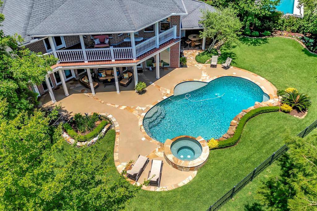 Private backyard featuring a resort style pool with a connected hot tub, surrounded by a stone patio and lush landscaping. The home has a covered patio with seating and an upper balcony, offering ample outdoor living space.