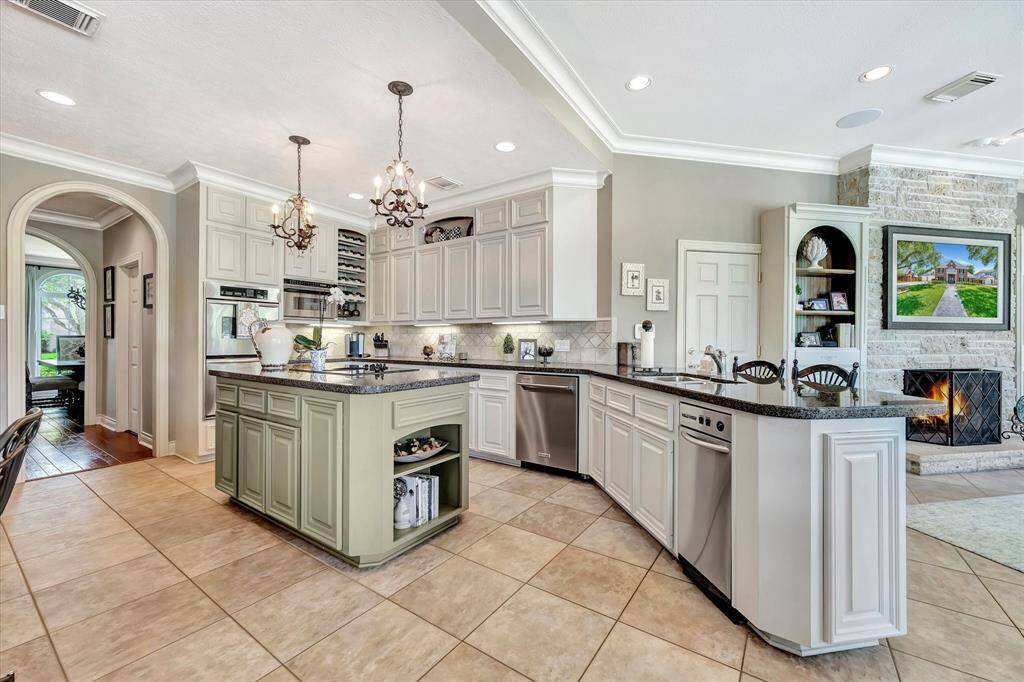 Island kitchen features elegant cabinetry, granite countertops, and a large breakfast bar with seating. It includes stainless steel appliances,and an open layout leading to a living area with fireplace perfect for entertaining. The tile flooring and tasteful lighting fixtures enhance the welcoming ambiance.