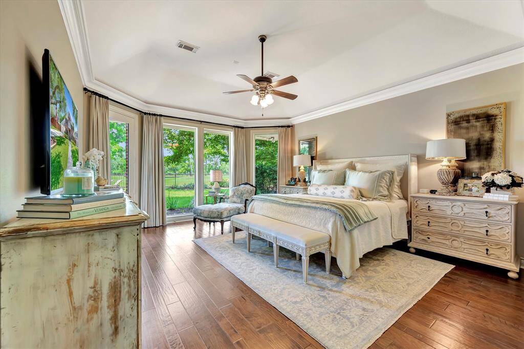 This primary bedroom features elegant hardwood flooring, a relaxing seating area, and large windows offering a scenic garden view. The room is well-lit with soft, natural light, complemented by tasteful decor, custom drapery and crown molding.