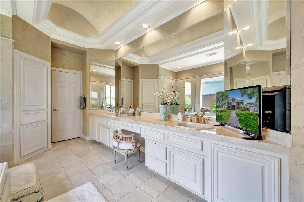 This luxurious bathroom features dual sinks with elegant countertops, ample mirror space, and soft recessed lighting. The neutral tones and decorative molding create a sophisticated ambiance, while a small TV adds modern convenience.